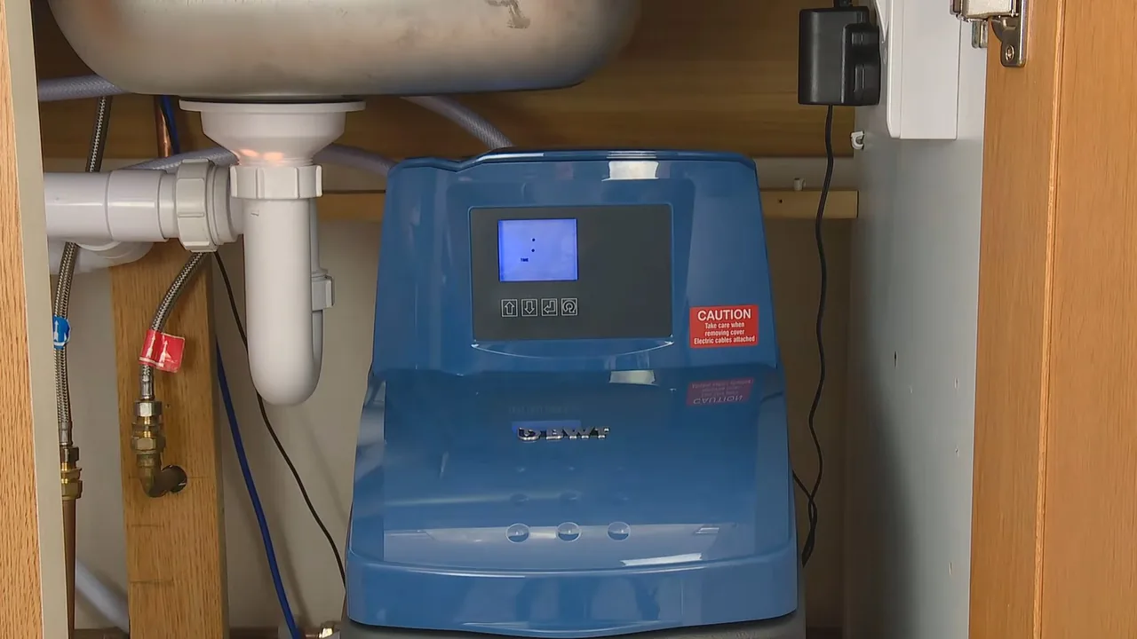 Blue water softener with a digital display standing under a kitchen sink beside the drain pipe.