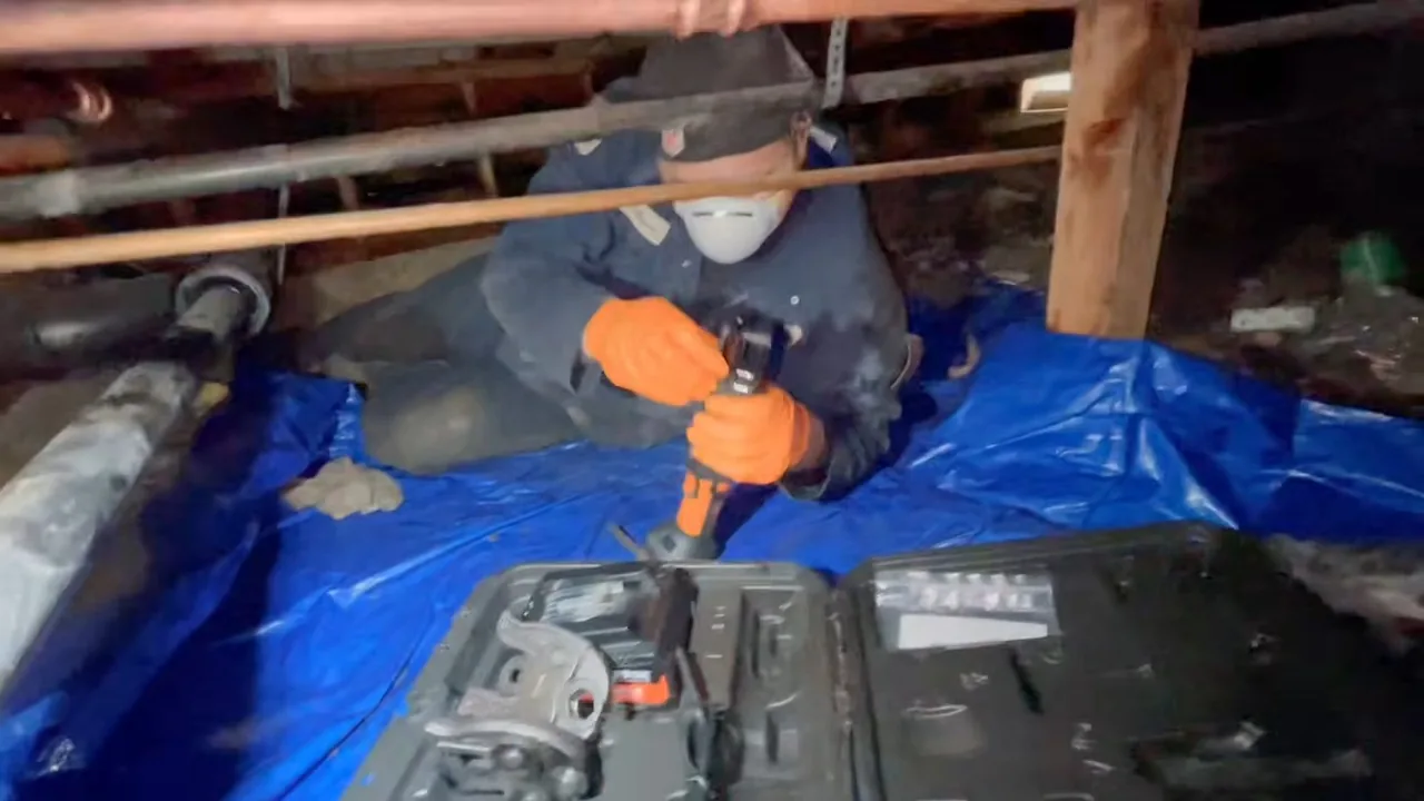 Worker in a respirator and orange gloves kneeling on a blue tarp in a crawlspace with a power tool and an open tool case, copper pipes overhead.