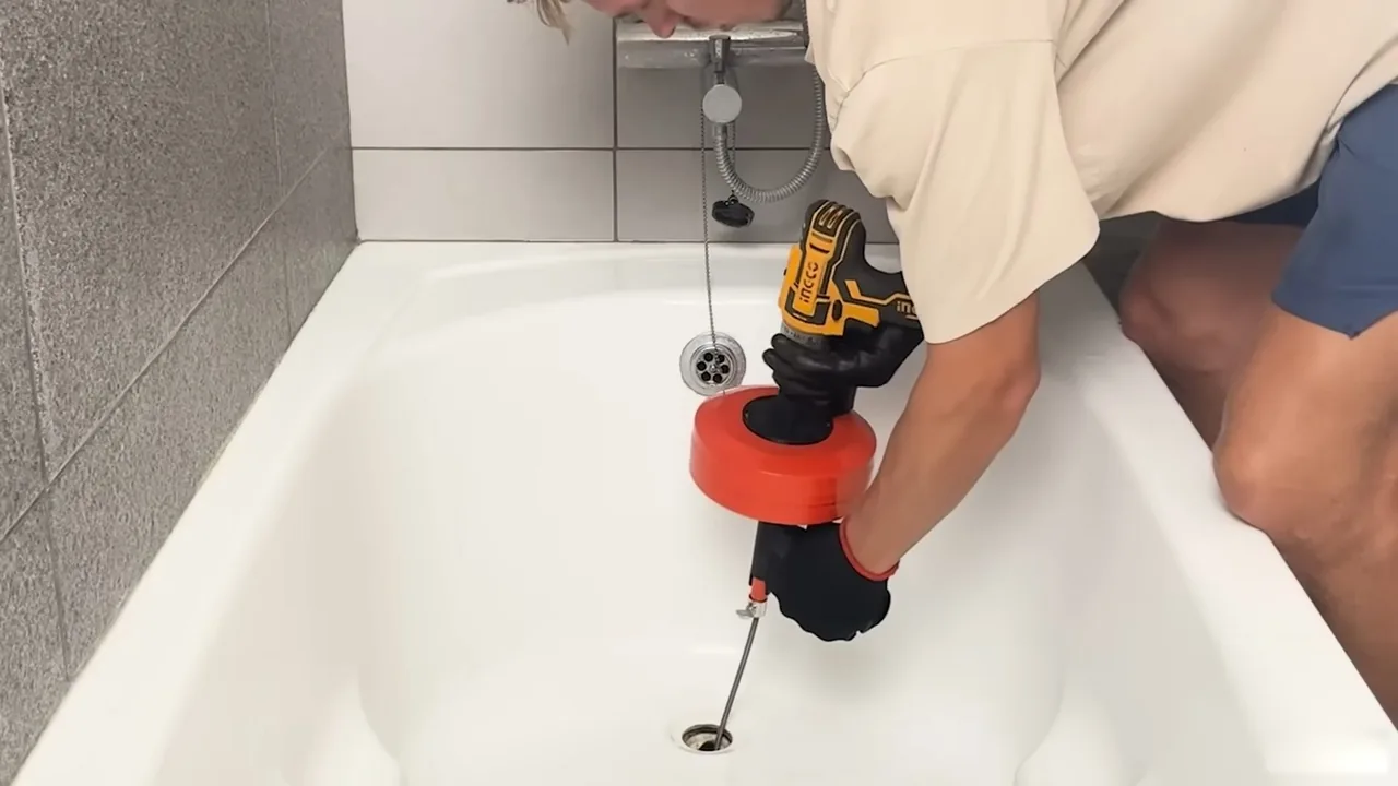 Plumber leaning over a bathtub to run a motorized drain auger down the tub drain.