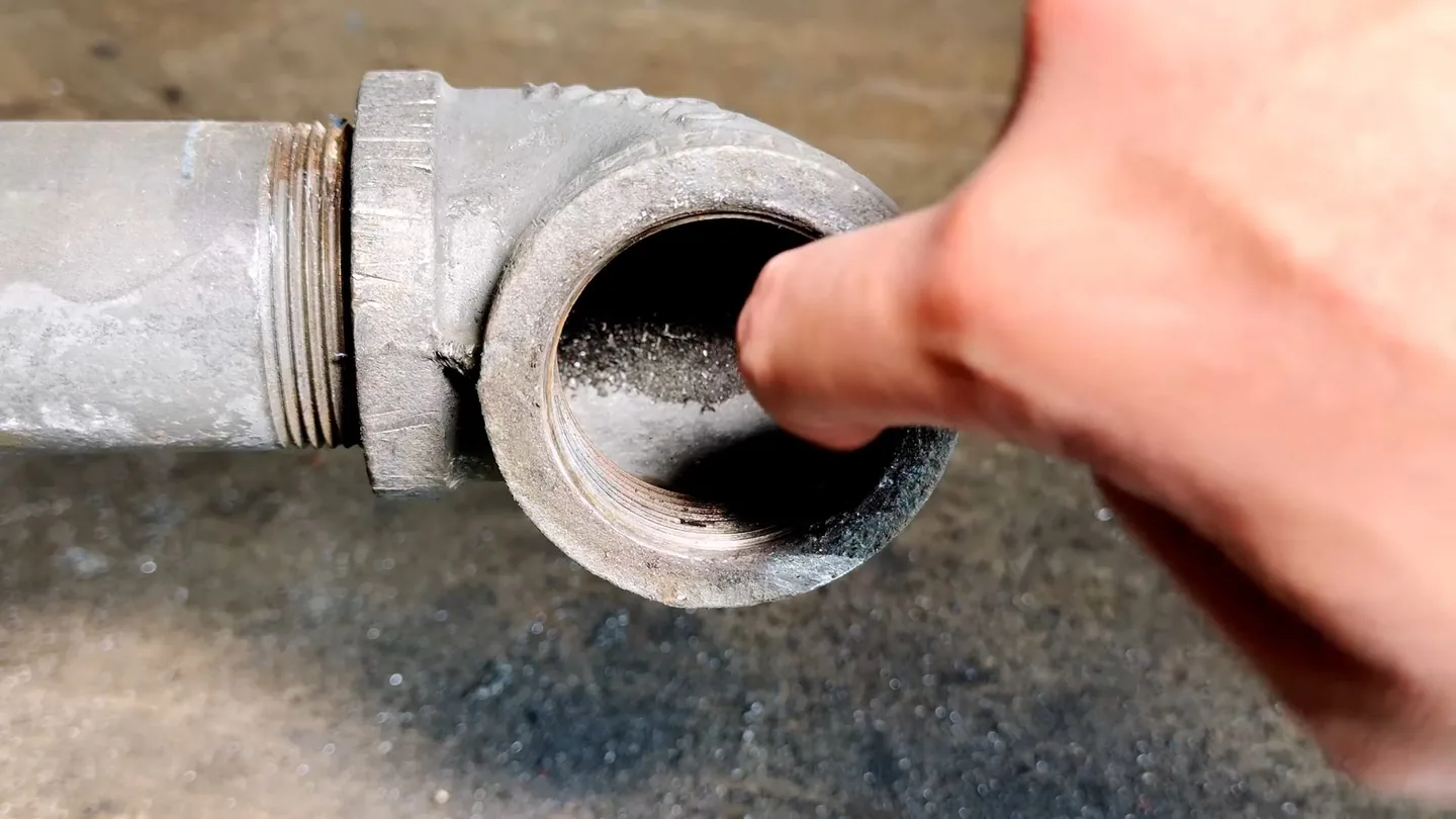 Finger pointing into the open end of a galvanized pipe elbow with its cap removed.