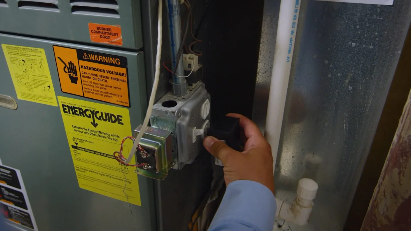Hand reaching to a gas control valve beside yellow warning and energy labels on a metal heater cabinet.