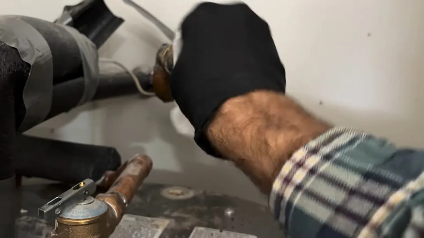 Gloved hand at a fitting on top of a water heater tank, with wrapped pipes behind and a brass valve below.