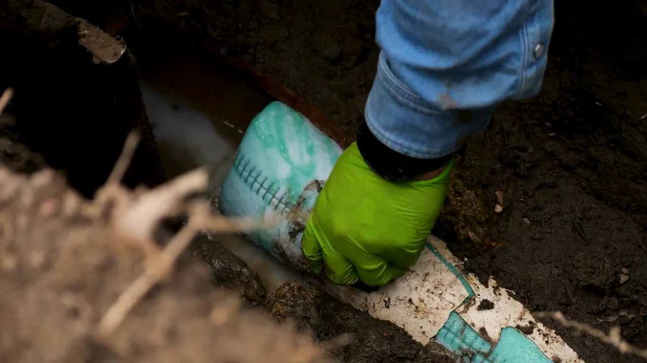 Gloved hand pressing a green-and-white lining tube against a pipe in a dirt trench.