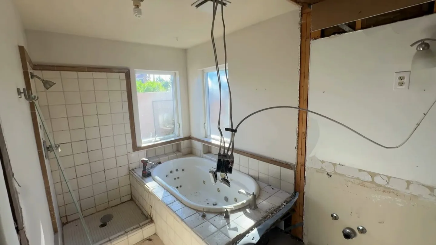 Gutted bathroom with open wall framing, a tiled tub deck and a light cable hanging from the ceiling.