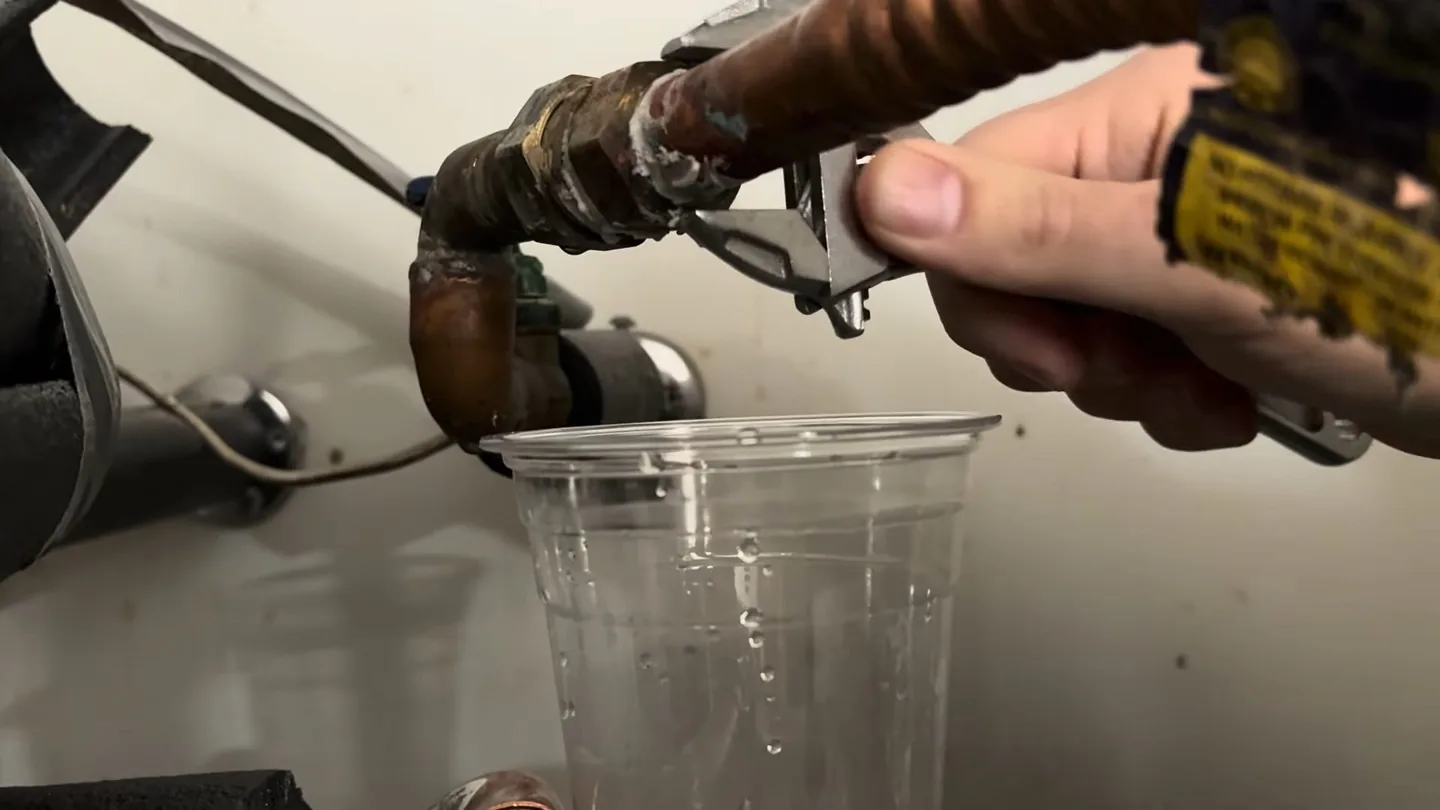 Hand gripping a pipe fitting on a water heater inlet with a clear cup catching drips beneath it.