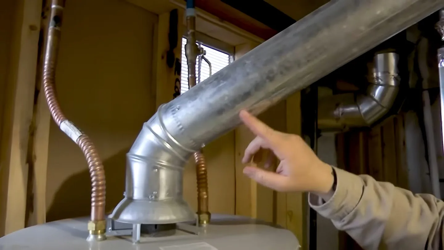 Hand pointing to a galvanized vent pipe where it meets the top of a water heater, with flexible copper lines nearby.