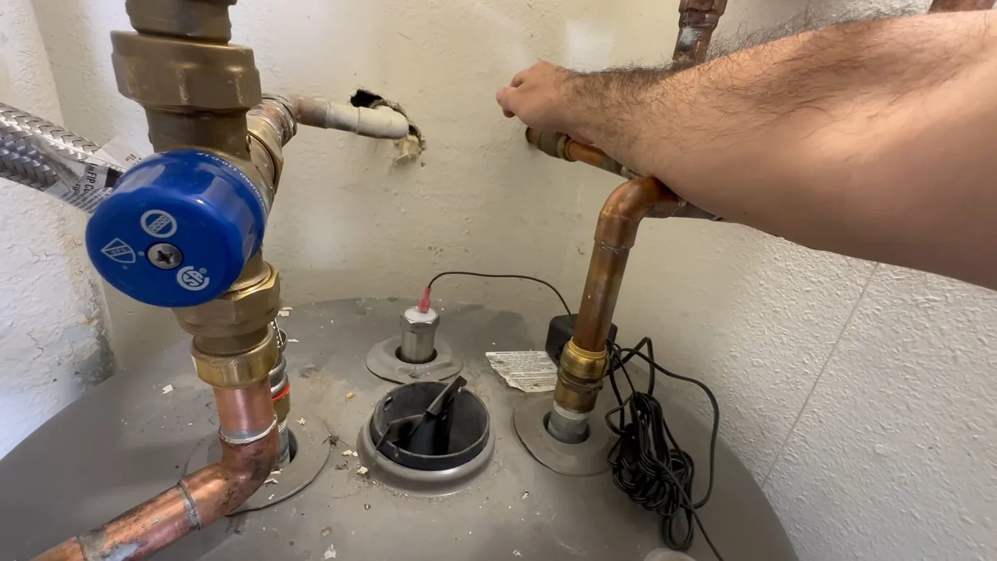 Hand reaching toward a blue-handled shutoff valve and copper pipes above a water heater tank.