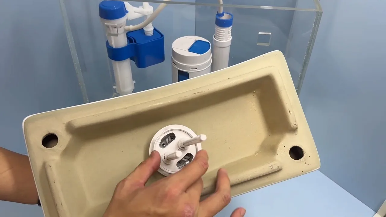 Hands working on the flush valve fitting at the bottom of a toilet tank, with a fill valve and cap assembly behind it.
