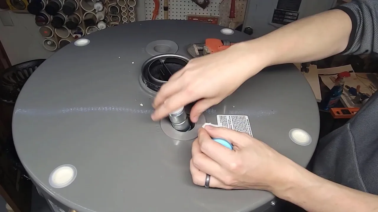 Hands working at a threaded opening on the top of a gray water heater tank, with a workbench behind.