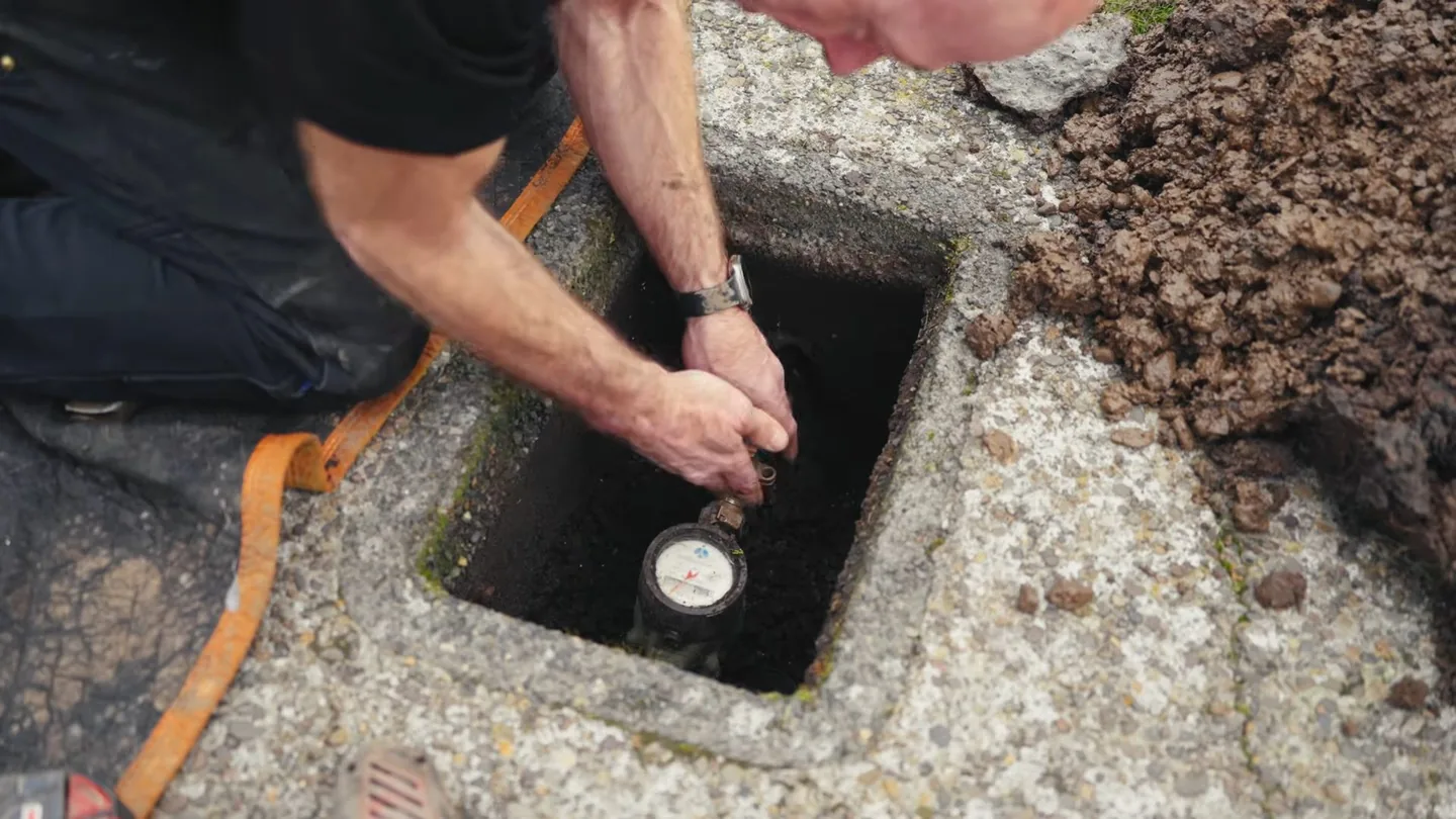 Man kneeling at an open water meter box in a paved area and reaching down toward the meter dial.