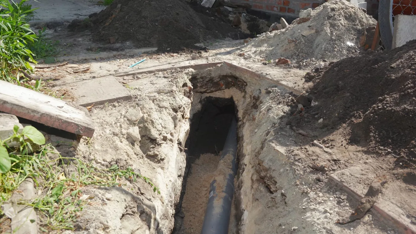 Narrow trench beside a house exposing a dark length of buried pipe, with piled soil and a board at the left.