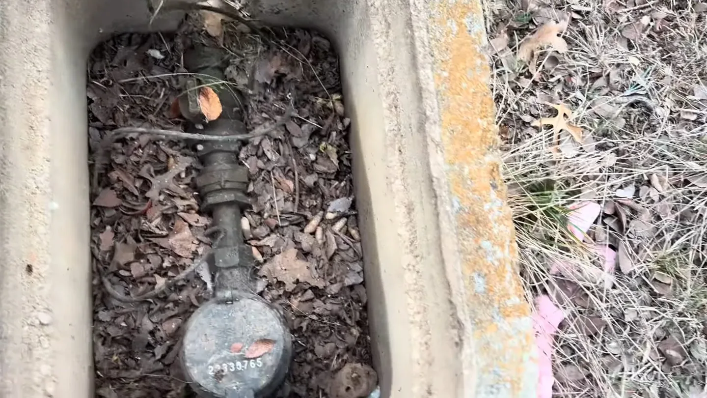 Open concrete meter box in a yard with a water meter and leaf litter around the pipe.