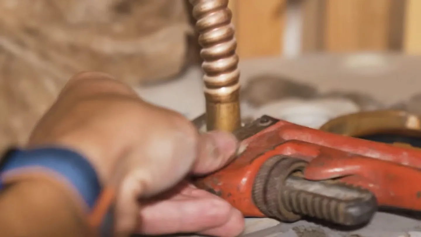 Hand holding an orange pipe wrench on a flexible copper supply line.