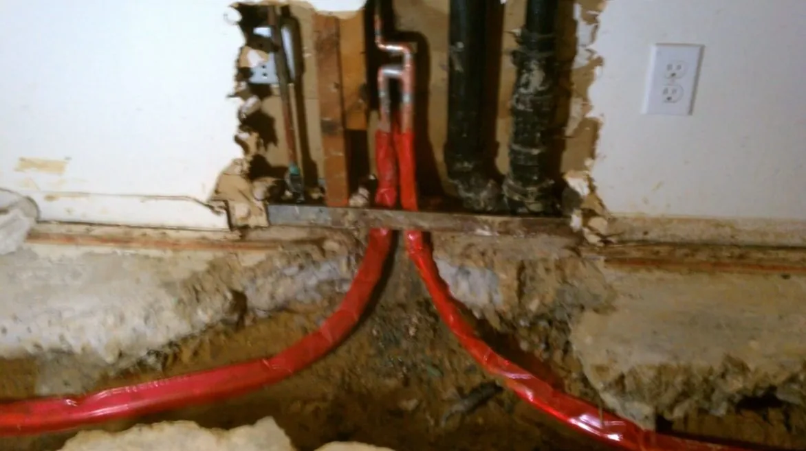 Cut-out section of a wall at floor level with red flexible tubing curving down from the opening and black pipe beside it.