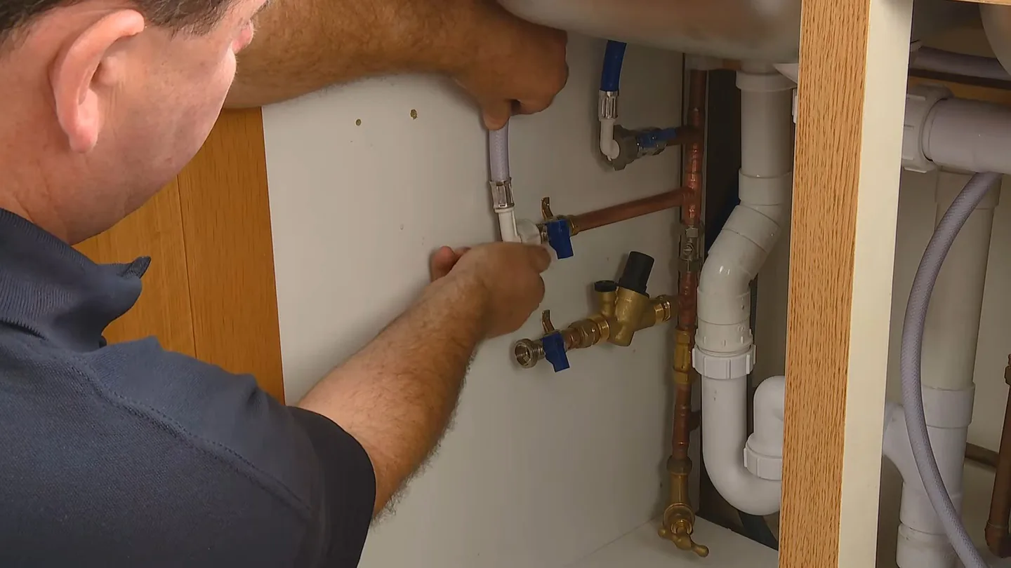 Technician reaching into an open sink cabinet to work on blue-handled shutoff valves, copper supply lines and a white drain trap.
