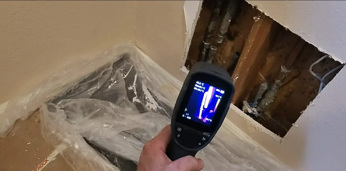 Thermal imaging camera held toward a cut opening in a corner wall, with plastic sheeting laid over the floor.