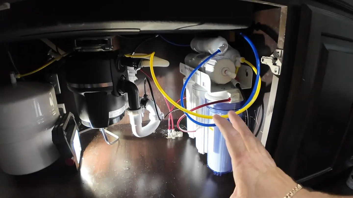 Open sink cabinet with a garbage disposal at the left, a white drain trap below it and tubing running to a filter system.