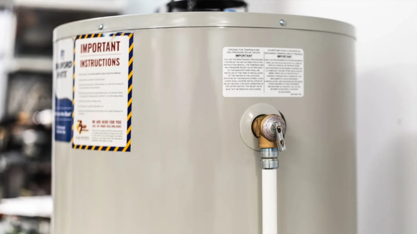 Top of a cream-colored tank water heater showing warning labels and a relief valve with a white line attached.