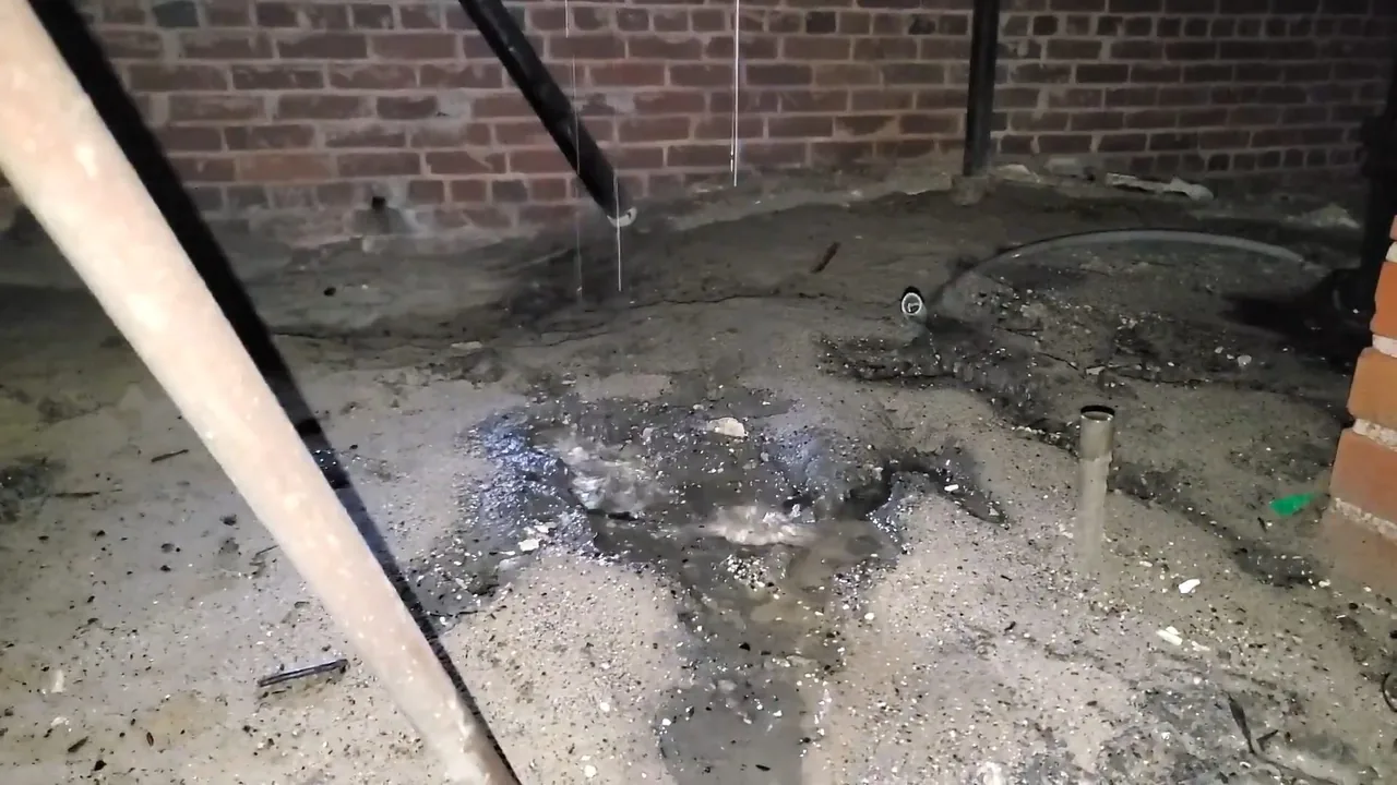 Wet concrete floor strewn with debris beside a brick wall, with a pipe leaning across the left side.